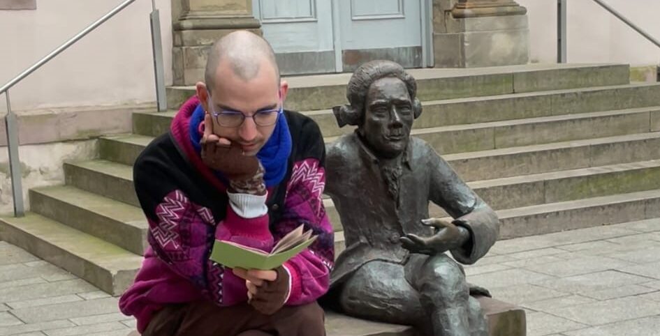 Philip Flacke sitting on a bench reading a pamphlet next to a statue of Lichtenberg